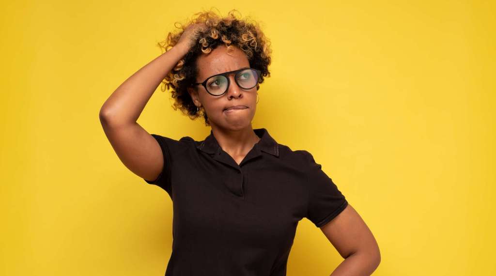Woman in glasses scratching her head against a yellow background, looking confused.