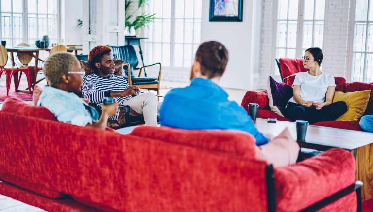 A diverse group of people engaged in a friendly conversation while sitting on red sofas in a bright, modern lounge.