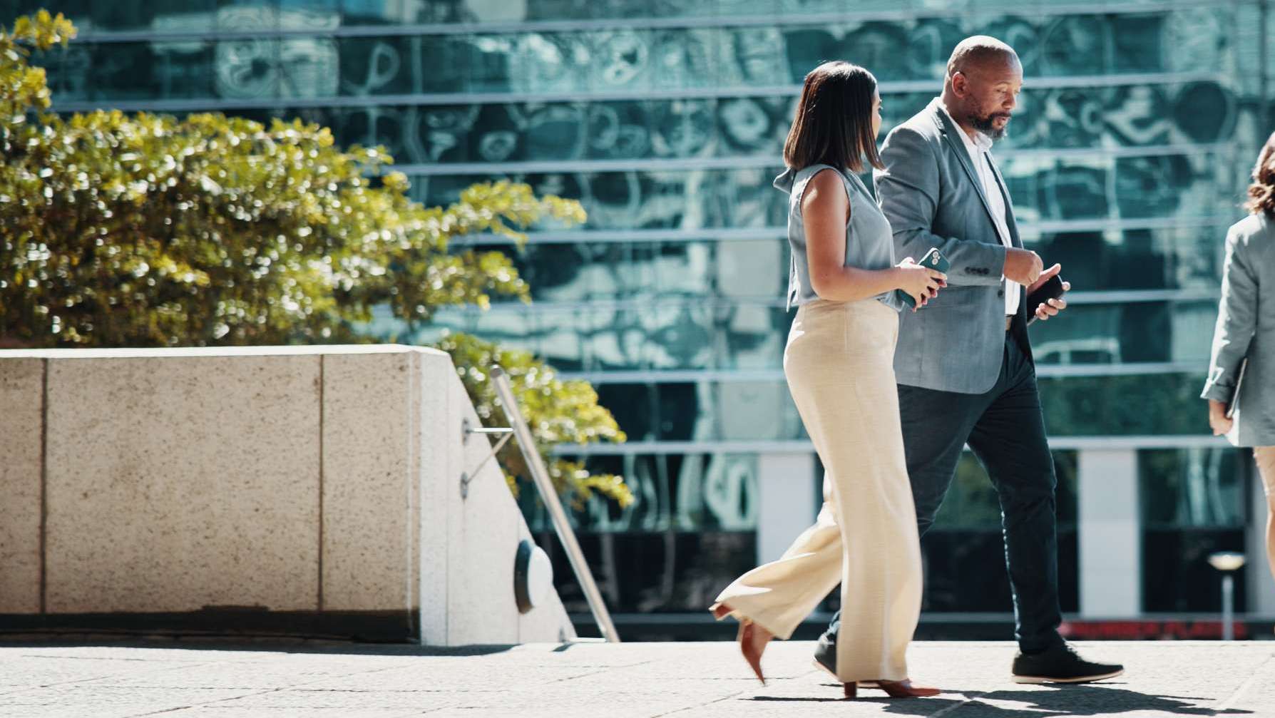 Two professionally dressed people walking outside a modern office building, engaged in conversation on a sunny day.