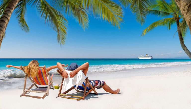 Couple relaxing in lounge chairs on a tropical beach under palm trees with a cruise ship in the distance.