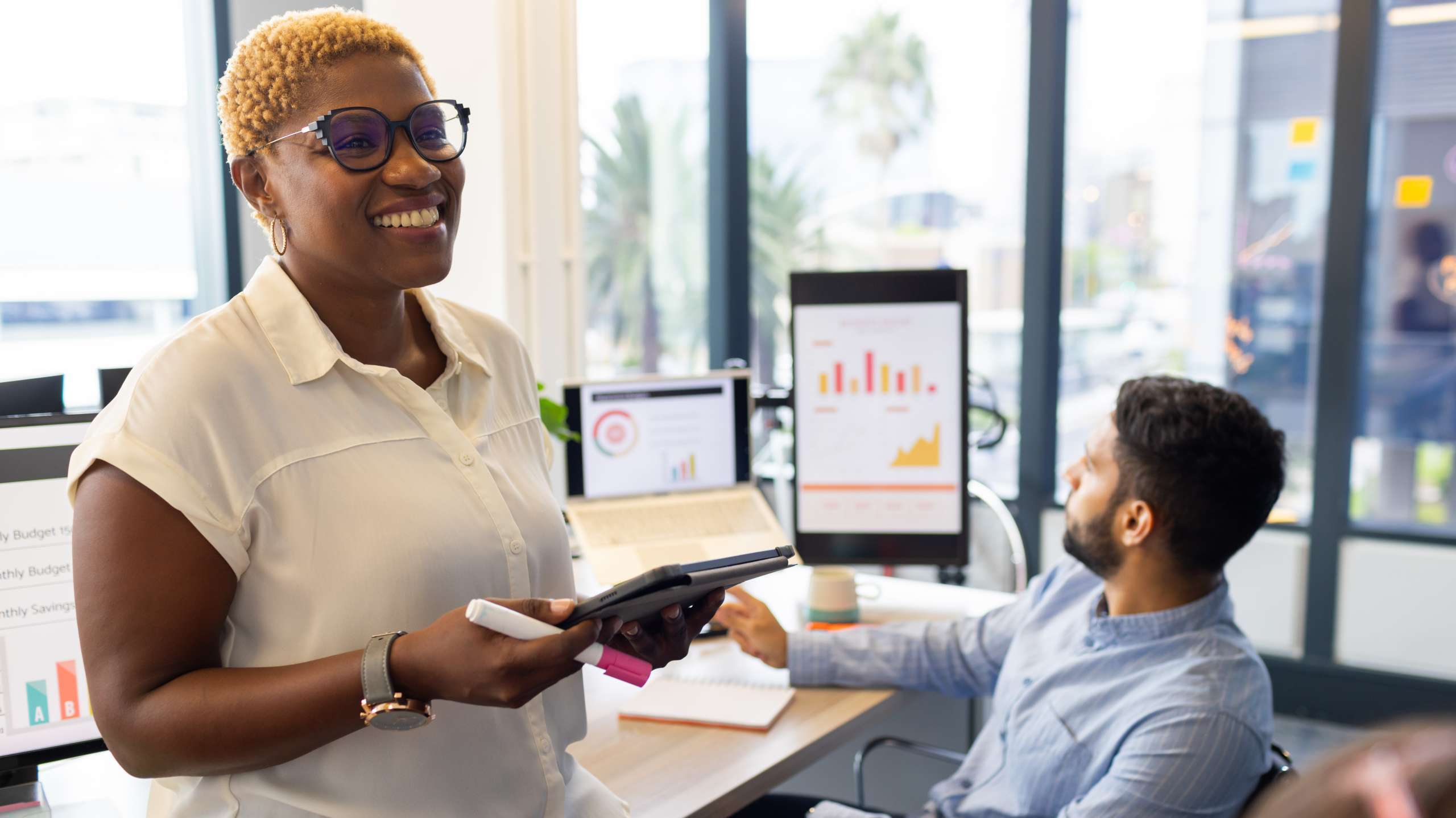 Smiling businesswoman holding a tablet and marker while presenting data to colleagues in a modern office with charts displayed on screens.