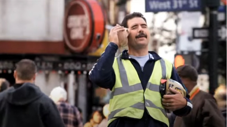 Man in a reflective safety vest smiling and holding a seashell to his ear like a phone while standing on a busy city street.