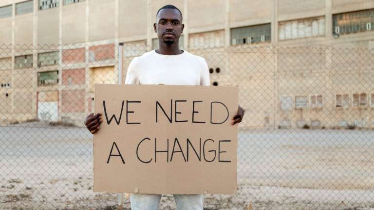 African American man holding a cardboard sign that says “We Need a Change,” symbolizing the need for church evolution and renewal.