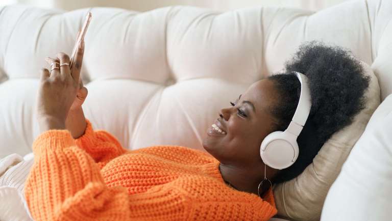 Smiling woman in an orange sweater with headphones, using her phone while relaxing on a couch — illustrating digital engagement in everyday life.