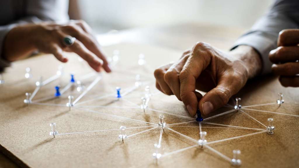 Hands placing pins connected with string on a corkboard, symbolizing a network of connections.