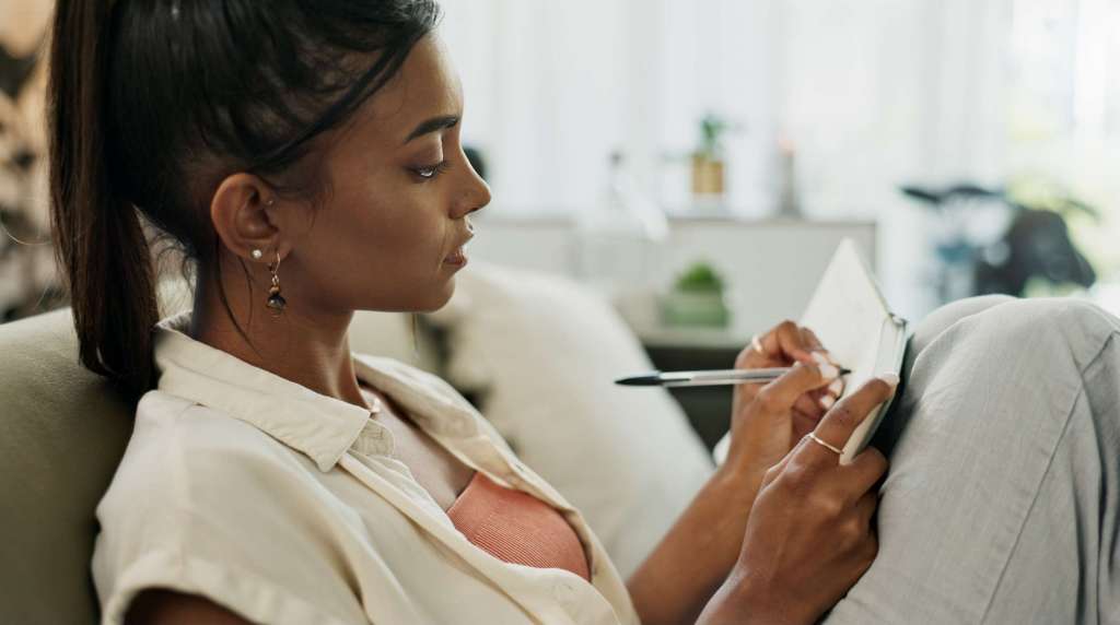 A woman thoughtfully writing in a notebook, seated comfortably indoors, symbolizing reflection, vision, and planning.