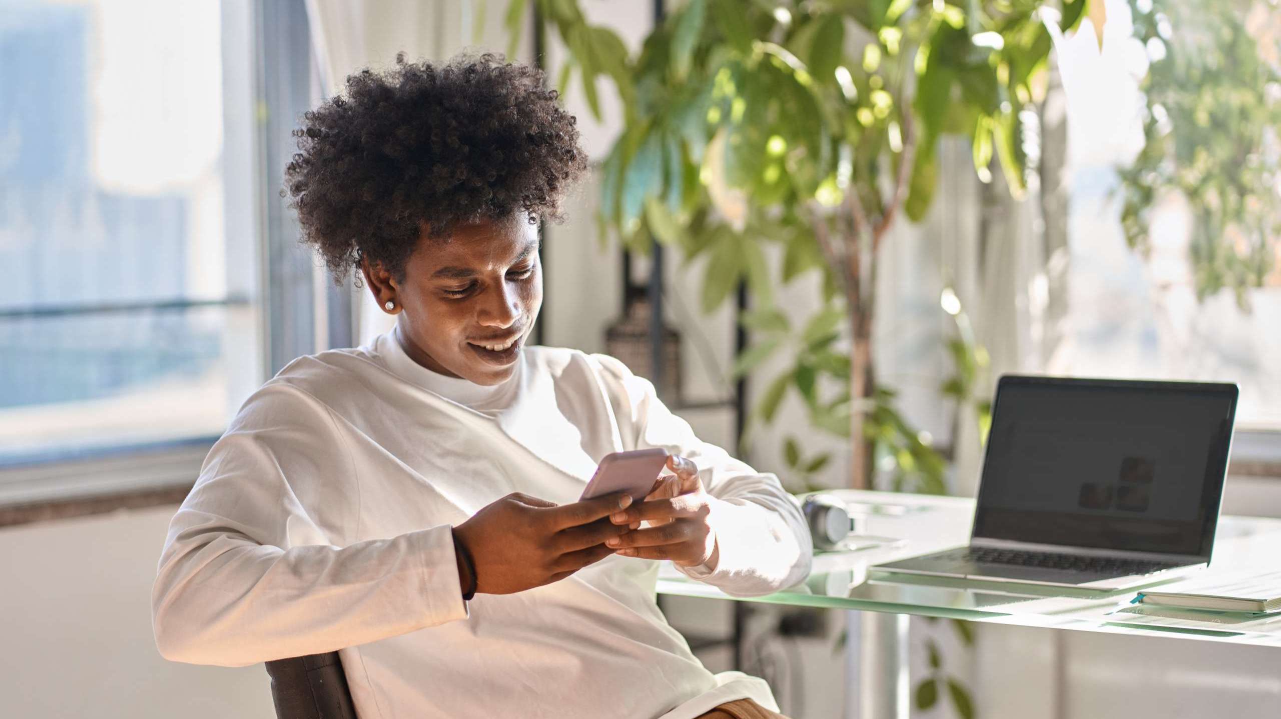 Young Black man smiling while using smartphone at a desk with laptop, representing social media engagement